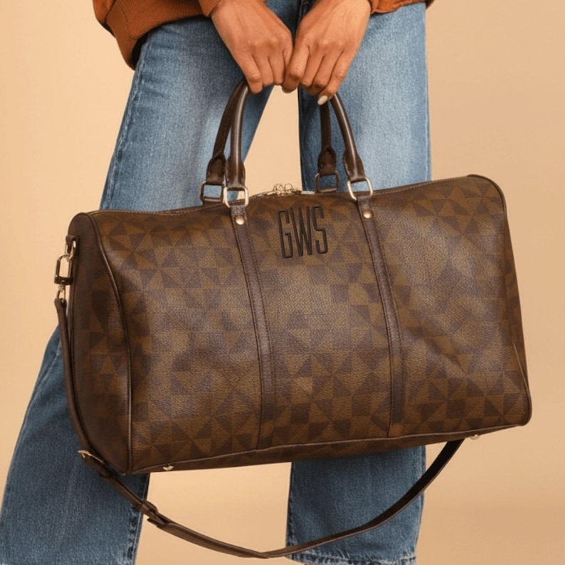 May include: A brown duffel bag with a geometric pattern and the initials "GWS" in the centre. The bag has two handles and a shoulder strap. The bag is held by a person wearing blue jeans.