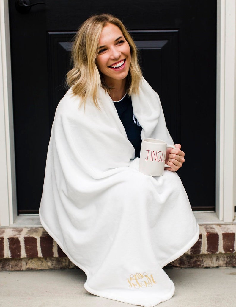 May include: A white fleece blanket with a gold monogram "KCM" embroidered on the bottom corner. A person is sitting on a brick step with the blanket wrapped around them, holding a white mug with the word "JINGLE" in red lettering.