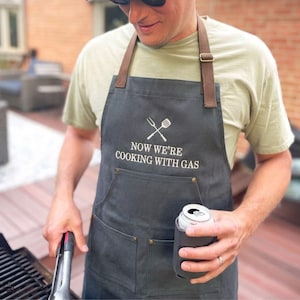 man outside grilling wearing a charcoal apron with custom embroidered text reading: "now we're cooking with gas"