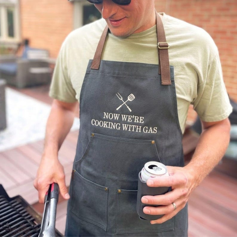 man outside grilling wearing a charcoal apron with custom embroidered text reading: "now we're cooking with gas"