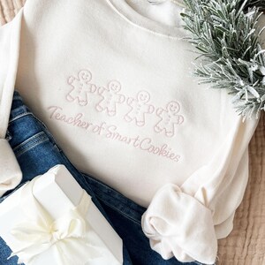 sweet cream crewneck sweatshirt with custom gingerbread men icons and "teacher of smart cookies" design embroidered in powder pink thread