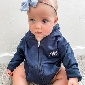baby girl wearing a full zip hooded navy sweatshirt with embroidered name on the left chest