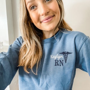 Op de afbeelding: Een blauw sweatshirt met de tekst "Kaylyn RN" en een geborduurd medisch symbool op de voorkant.