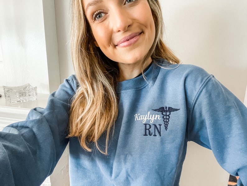 Op de afbeelding: Een blauw sweatshirt met de tekst "Kaylyn RN" en een geborduurd medisch symbool op de voorkant.