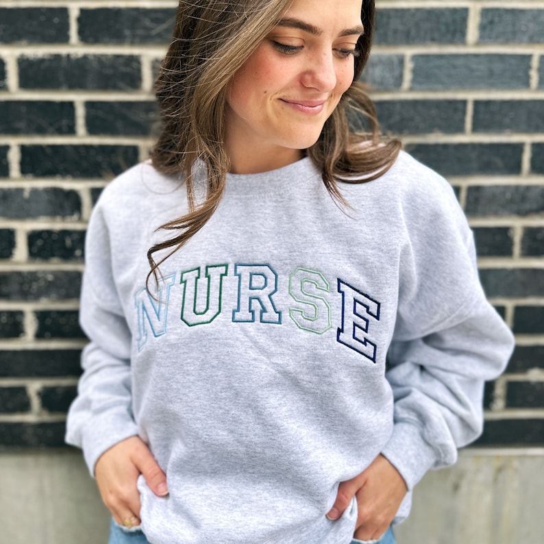 young woman wearing an ash crewneck sweatshirt with custom NURSE embroidered design in shades of blue and green