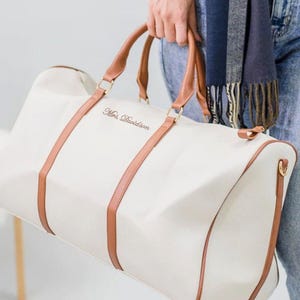 May include: Cream duffel bag with brown leather handles and accents. The bag has the text "Mrs. Davidson" in a script font. The bag is held by a person wearing blue jeans and a scarf.