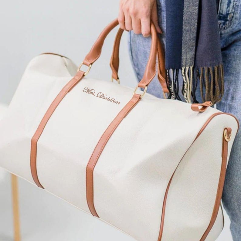 May include: Cream duffel bag with brown leather handles and accents. The bag has the text "Mrs. Davidson" in a script font. The bag is held by a person wearing blue jeans and a scarf.