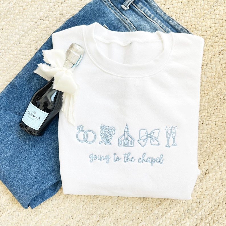 May include: A white sweatshirt with light blue embroidered wedding-themed icons and text that reads "going to the chapel." A small bottle of sparkling wine with a white bow is next to the sweatshirt. The sweatshirt is on top of blue jeans.