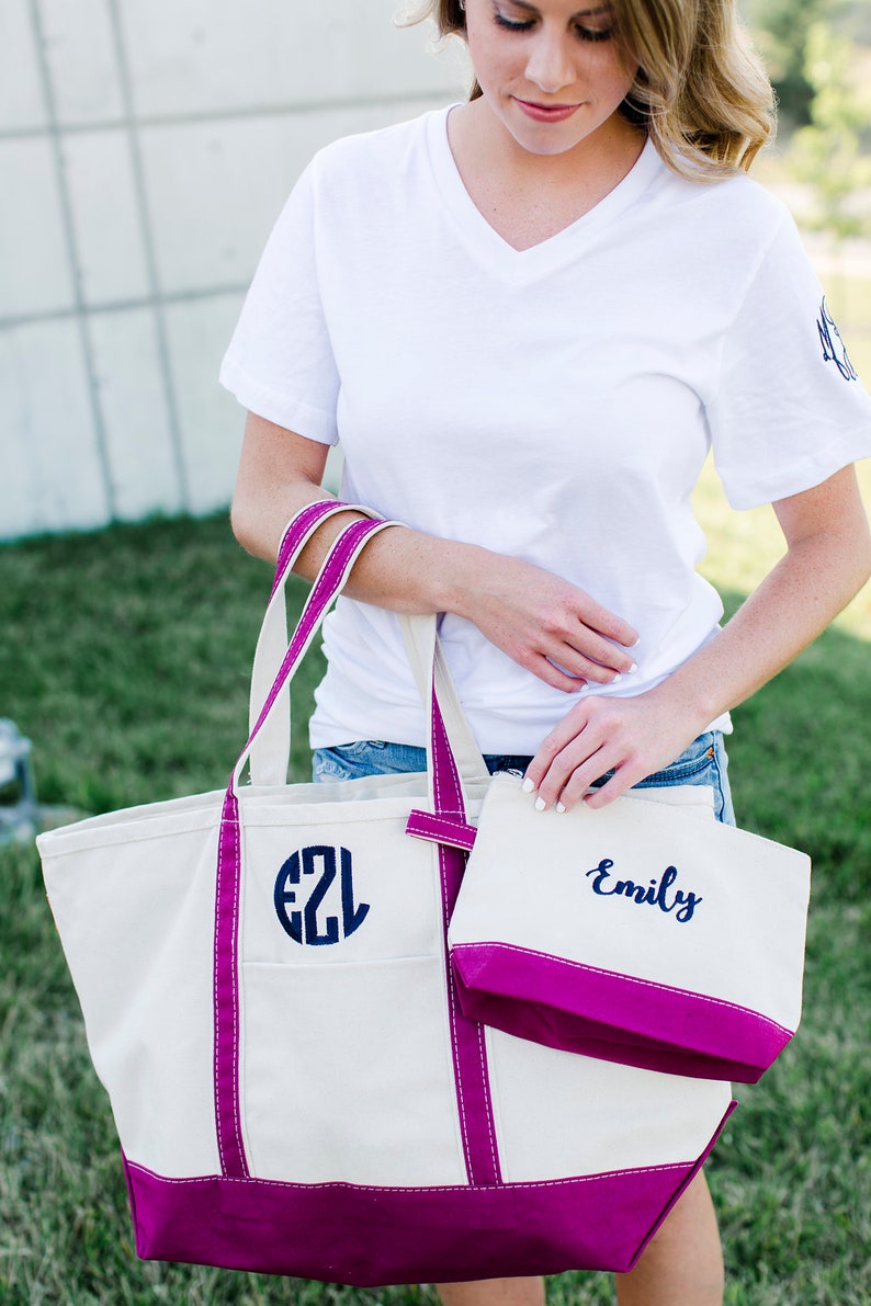 Pode incluir: Uma bolsa tote de lona branca com acabamento rosa e um monograma azul marinho "EZL" na frente. Uma bolsa de lona branca menor com acabamento rosa e o nome "Emily" bordado em azul marinho est&aacute; presa &agrave; bolsa tote.