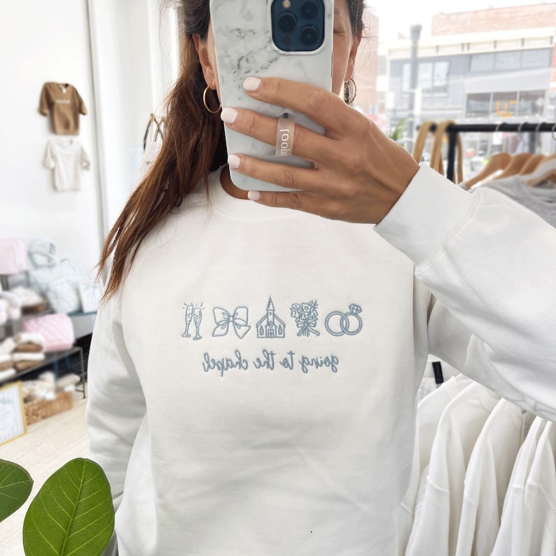 May include: White sweatshirt with light blue embroidered wedding-themed icons and text. The design includes champagne glasses, a bow, a church, flowers, and rings. The text reads "Save the date" in a script font.