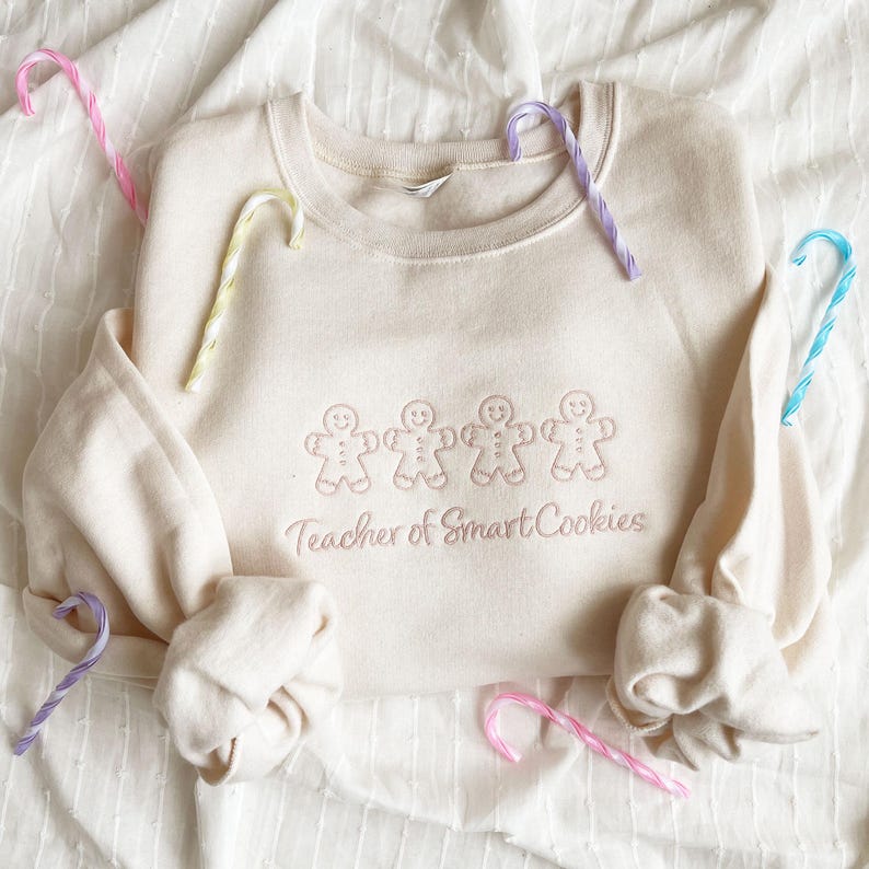 sweet cream crewneck sweatshirt with custom gingerbread men icons and "teacher of smart cookies" design embroidered in powder pink thread