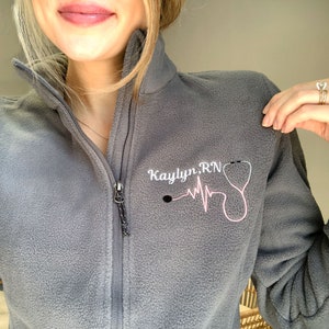 Peut inclure: Veste en polaire grise avec fermeture &eacute;clair et un motif brod&eacute; rose qui dit "Kaylyn.RN" avec un st&eacute;thoscope et un graphique de battement de c&oelig;ur.