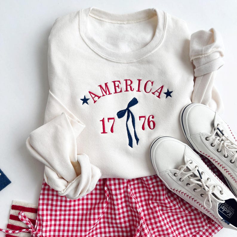 May include: Cream-colored sweatshirt with the word "AMERICA" embroidered in red, along with the number "1776" and a blue bow. Paired with red and white checkered shorts and white sneakers.