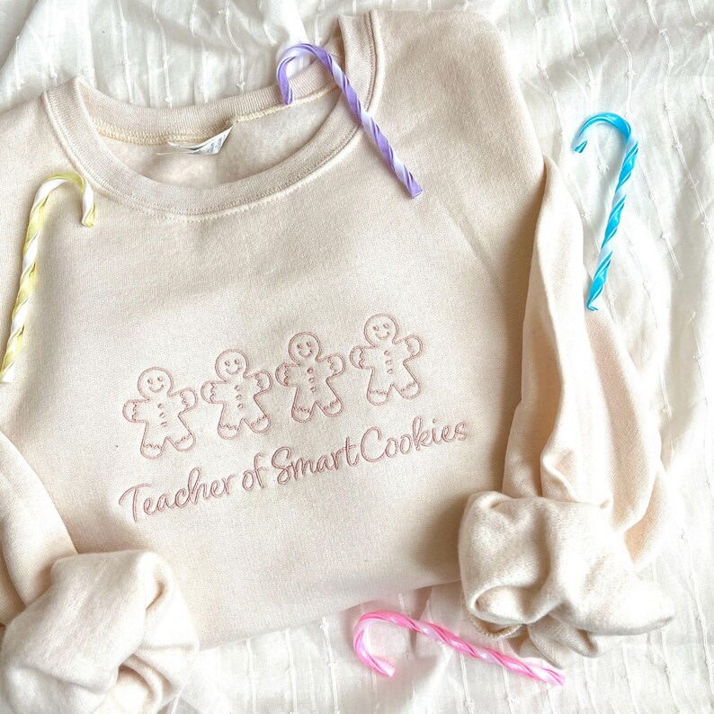 sweet cream crewneck sweatshirt with custom gingerbread men icons and "teacher of smart cookies" design embroidered in powder pink thread