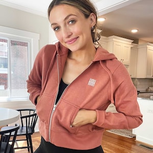 May include: A rust-coloured cropped zip-up hoodie with a hood and a small pocket on the left side. The hoodie has the letters "KAH" embroidered on the front.