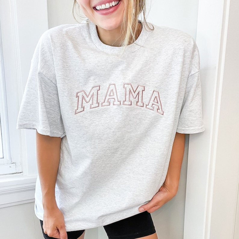 K&ouml;nnte beinhalten: Ein graues Oversize-T-Shirt mit dem Wort "MAMA" in rosa gestickten Buchstaben.