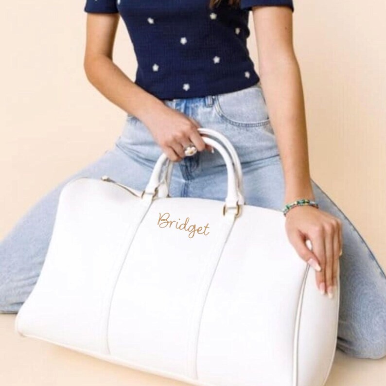 May include: White duffel bag with gold-coloured hardware and the name "Bridget" in gold script. The bag has two handles and a zip closure. The bag is made of a smooth material and is a neutral colour.