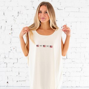 blonde woman standing in front of a white brick wall wearing denim shorts and an oversized ivory comfort colors tank top with embroidered mini usa flags and dainty bow design across the chest in an alternating pattern
