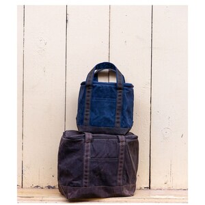 May include: Two canvas tote bags stacked on top of each other. The top bag is dark blue with black straps and the bottom bag is brown with black straps.