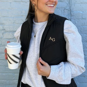 May include: A black quilted vest with a gold zipper and the initials "AG" embroidered on the front. The vest is worn over a gray long-sleeved shirt.