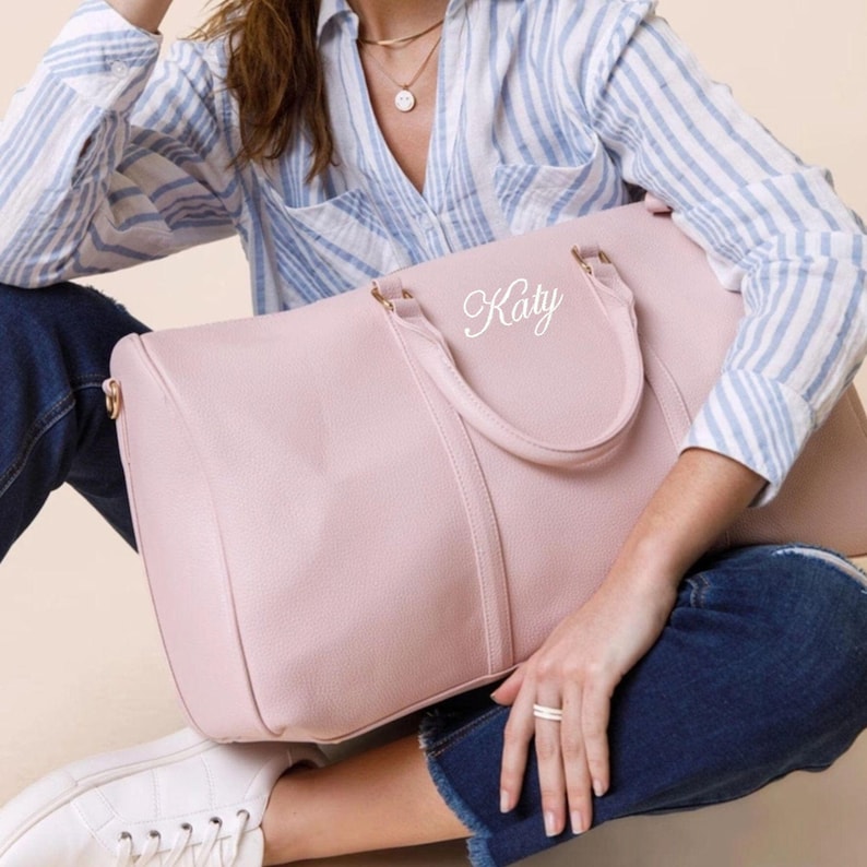 May include: A pale pink duffel bag with gold-coloured hardware and handles. The bag has the name "Katy" in white script. The bag is on a person's lap, who is wearing blue jeans, a striped shirt, and white trainers.