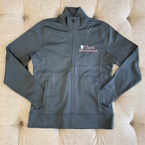 Puede incluir: Una chaqueta gris oscuro con cremallera que tiene un peque&ntilde;o logotipo bordado que dice "Clara Dental Assistant" y un icono de diente.