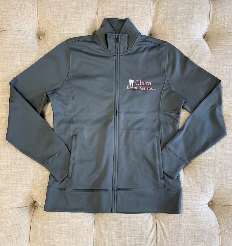 Puede incluir: Una chaqueta gris oscuro con cremallera que tiene un peque&ntilde;o logotipo bordado que dice "Clara Dental Assistant" y un icono de diente.
