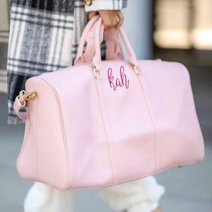 May include: A pale pink duffel bag with gold-coloured hardware and the initials "kah" in a cursive font. The bag has two handles and a shoulder strap. The person holding the bag is wearing a black and white plaid coat.