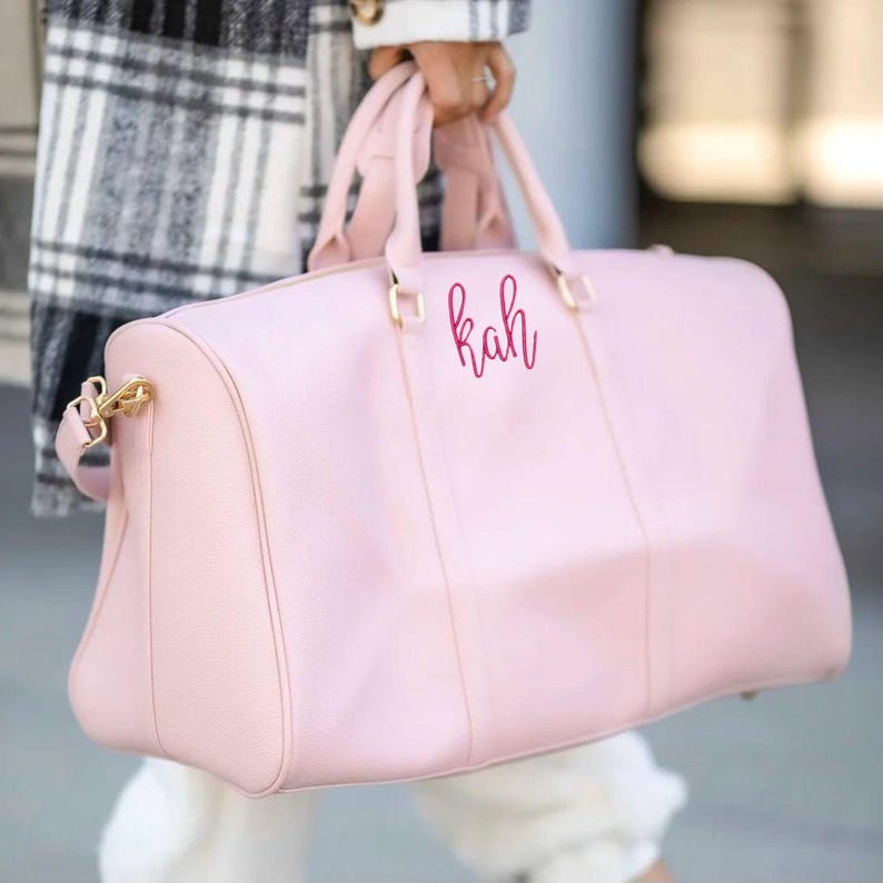 May include: A pale pink duffel bag with gold-coloured hardware and the initials "kah" in a cursive font. The bag has two handles and a shoulder strap. The person holding the bag is wearing a black and white plaid coat.