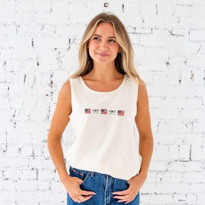 blonde woman standing in front of a white brick wall wearing denim shorts and an ivory comfort colors tank top with embroidered mini usa flags and dainty bow design across the chest in an alternating pattern