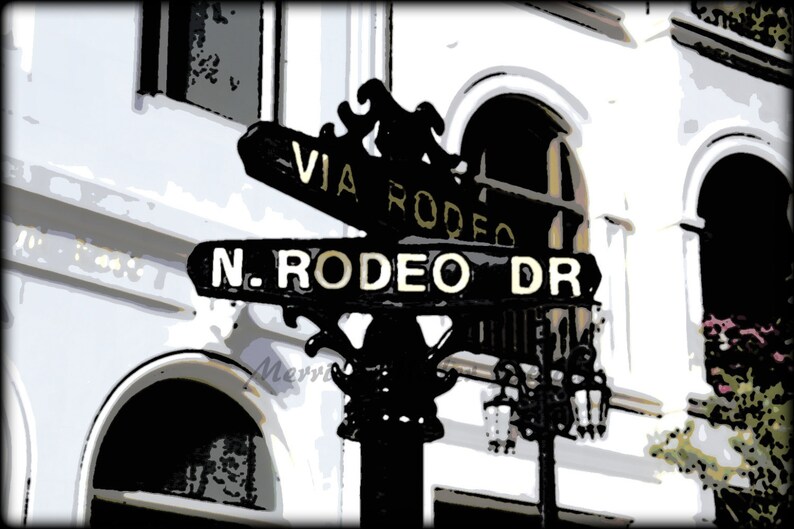 May include: Black and white street sign with gold lettering that reads "Via Rodeo" and "N. Rodeo Dr."