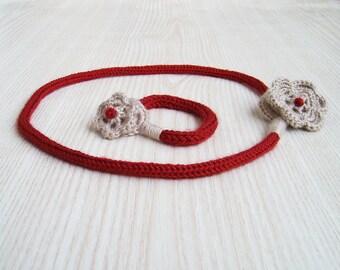 May include: A red crocheted necklace with two beige crocheted flowers, each with a red center. The necklace is lying on a white surface.
