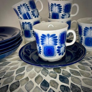 May include: A collection of white ceramic mugs, saucers, and bowls. The mugs and bowls have a dark blue floral pattern. The saucers and bowls are a solid dark blue color. The set is arranged on a patterned surface.