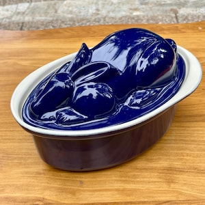 May include: A deep blue ceramic baking dish with a matching lid shaped like a resting rabbit. The oval-shaped base features a white rim, while the glossy, dark blue lid depicts the sculpted form of a rabbit in a curled, sleeping position.