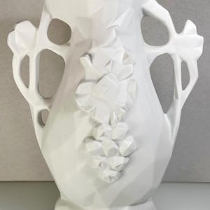 May include: A white ceramic vase with a geometric design. The vase features a grape cluster and leaf motif, with handles on either side. The vase is approximately 30 cm tall.