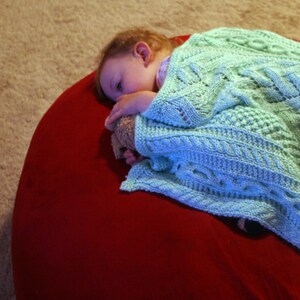 May include: A young child is sleeping on a red beanbag chair, covered with a light blue knitted blanket. The child is holding a stuffed animal.