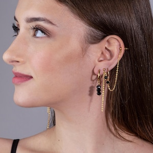 Puede incluir: Aretes de aro dorados con una cadena colgante y cuentas negras. La cadena está unida a un arete de barra dorado.