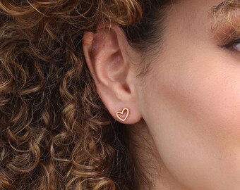 Puede incluir: Primer plano de una oreja con un pendiente de botón dorado en forma de corazón. El pendiente es un simple contorno de un corazón. La persona tiene el pelo castaño rizado y la piel clara.