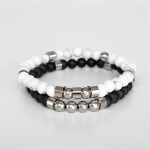 May include: Two bracelets, one with white and black beads and the other with silver beads. The bracelets are stacked on top of each other.