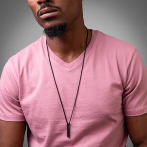 Pode incluir: Um colar de corrente preto com um pingente preto longo e fino pendurado no pescoço de uma pessoa. A pessoa está vestindo uma camiseta rosa de gola em V.