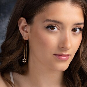 Puede incluir: Pendientes colgantes dorados con un colgante de estrella de David. Los pendientes tienen una delicada cadena que cuelga de la oreja, con un pequeño dije calado de estrella de David en la parte inferior.