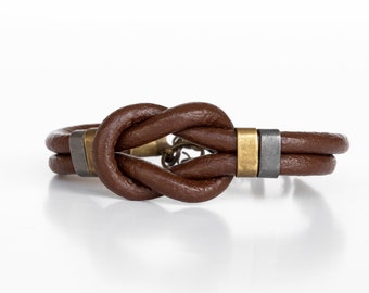 May include: A brown leather bracelet with a knotted design and metal accents. The bracelet has a gold and silver finish.