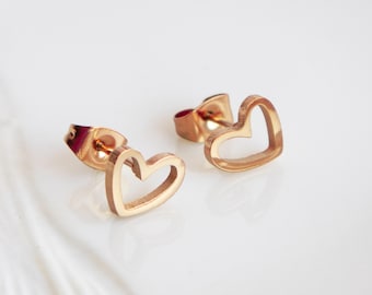 Puede incluir: Un par de pendientes de botón dorados con forma de corazón. Los pendientes son de metal brillante y tienen un diseño sencillo y abierto. La forma de corazón está claramente definida. Los pendientes se muestran sobre una superficie blanca.