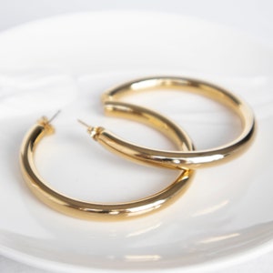 Könnte beinhalten: Ein Paar goldfarbene Creolen. Die Ohrringe sind dick und haben eine glatte, glänzende Oberfläche. Sie liegen auf einer weißen, leicht strukturierten Oberfläche. Ein klassisches Schmuckstück.