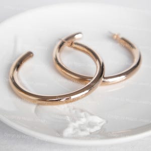 Könnte beinhalten: Ein Paar goldfarbene Creolen. Die Ohrringe sind rund und haben eine glatte, polierte Oberfläche. Sie liegen auf einer weißen, leicht strukturierten Oberfläche. Die Ohrringe haben einen warmen, metallischen Farbton.