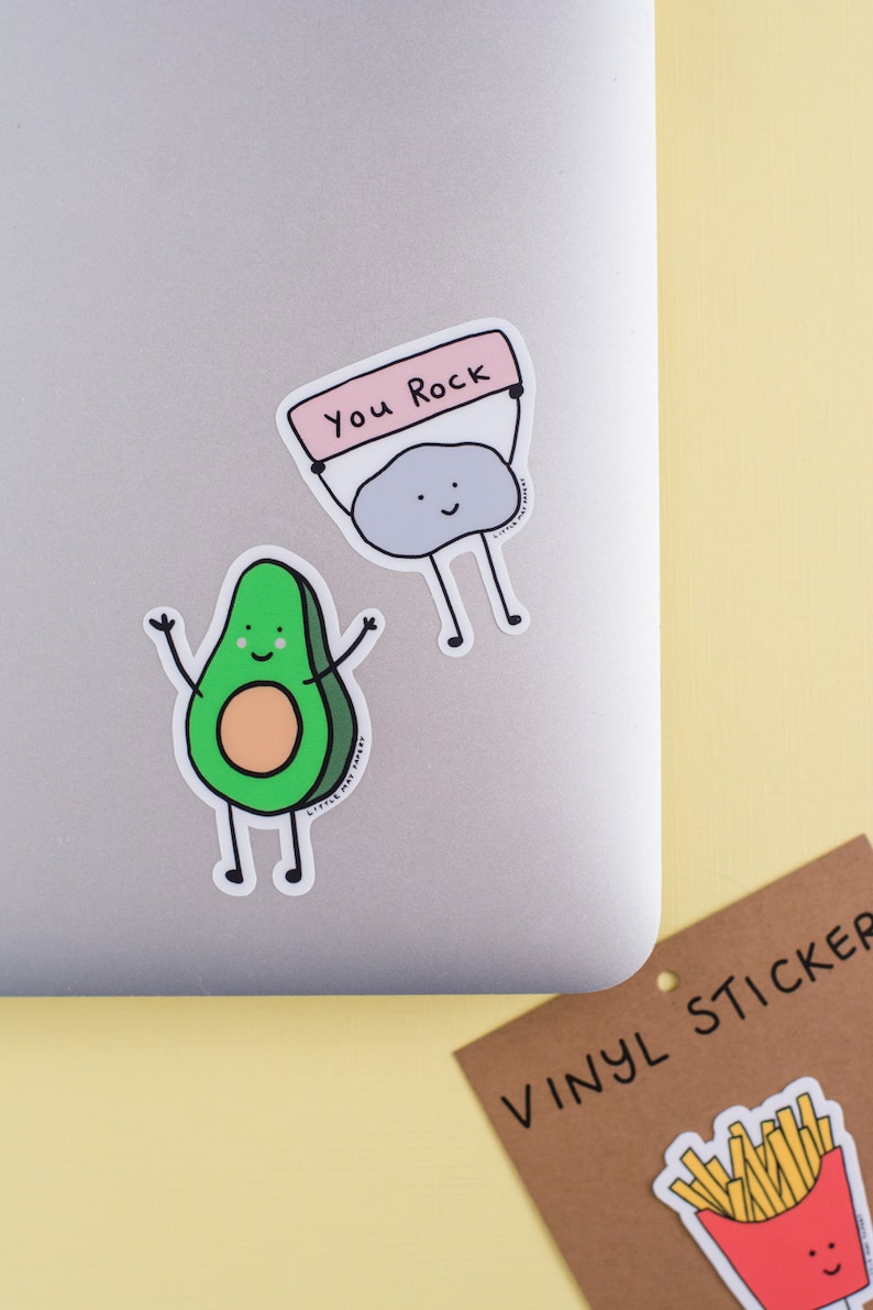 May include: Two vinyl stickers on a laptop. One is a green avocado with arms raised. The other is a gray rock holding a sign that says "You Rock". A third sticker of french fries is on a brown card that says "VINYL STICKER".