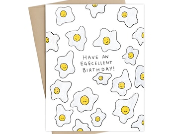 Może przedstawiać: Biała kartka z czarno-żółtą ilustracją jajka sadzonego z uśmiechniętą buźką. Tekst "HAVE AN EGGCELLENT BIRTHDAY!" jest napisany czarnym kolorem na kartce.