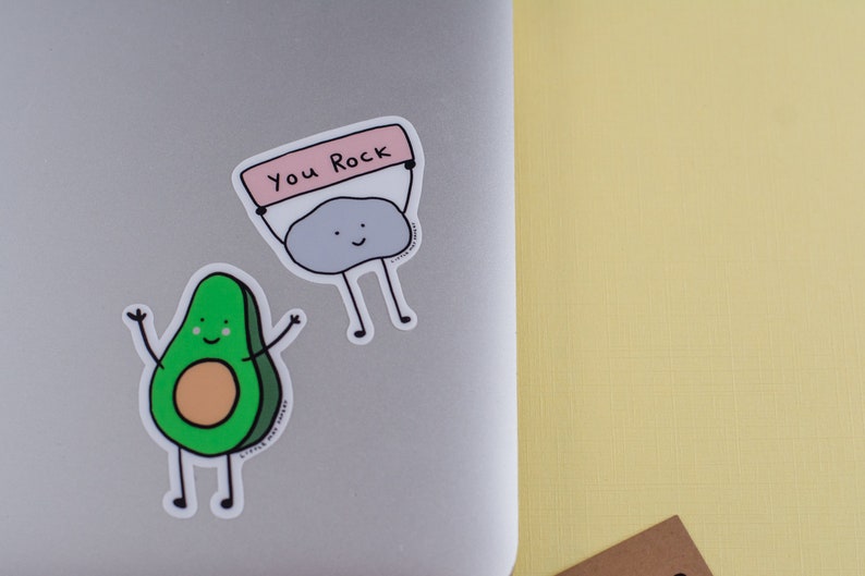 May include: Two stickers on a laptop. One is a green avocado with a smiling face and arms raised. The other is a gray rock holding a sign that says "You Rock". The background is a light yellow surface.