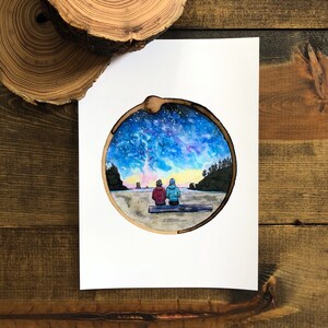 May include: A watercolor painting of two people sitting on a log, looking out at a starry night sky. The painting is framed in a round wooden circle.