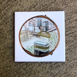 Pode incluir: Uma ilustra&ccedil;&atilde;o em aquarela de um horizonte da cidade com uma ponte e um bonde. A imagem &eacute; emoldurada em um c&iacute;rculo com uma borda marrom.